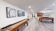Located at 2900 Pharr Court South NW in Atlanta's Buckhead neighborhood, this single-floor amenity space offers a thoughtfully designed layout ideal for socializing and recreation. Light gray wood-look flooring flows seamlessly throughout, unifying the open floor plan under recessed lighting and crisp white walls accented with crown molding.

The expansive game room serves as a centerpiece, equipped with a pool table and shuffleboard table alongside comfortable seating areas with modern teal and yellow chairs. Atlanta-themed artwork adorns the walls, adding a local character to the space. Natural light filters in through glass entry doors, enhancing the bright and welcoming atmosphere.

The living room features a tan leather sofa with teal accent pillows, a wall-mounted flat-screen TV, and a sleek tile-surround fireplace. Large sliding windows offer views to an outdoor area, while a glass-paneled door connects to an adjacent meeting room with a chandelier and conference-style seating.

The kitchen is finished with white shaker cabinetry, quartz countertops, a mosaic tile backsplash accent, a built-in microwave, and a matte black faucet over an undermount sink. A large island with a warm wood base provides additional prep and gathering space, illuminated by a modern pendant light. The overall ambiance is clean, contemporary, and community-focused.