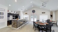 Located in Kingsport, Tennessee, this single-story space blends rustic charm with modern design throughout its thoughtfully arranged layout. The main living area immediately draws attention with its soaring vaulted ceilings, recessed lighting, and a dramatic floor-to-ceiling stacked stone fireplace with a wood mantel — a true centerpiece of the room. Comfortable seating surrounds a stylish coffee table, making this an inviting gathering space. Adjacent to the living area, an open-concept kitchen and bar area features dark cabinetry, a light countertop surface, and a reclaimed wood-paneled island with bar stool seating. French doors flood the space with natural light and provide direct access to the outdoors. A dedicated dining and activity area offers round table seating alongside decorative arrow wall accents and a workspace station tucked along the windowed wall. Throughout the space, neutral carpet flooring, white walls, and ceiling fans contribute to a clean, airy atmosphere. Glass-paned French doors connect distinct zones, maintaining an open feel while defining each area. The cohesive rustic-modern aesthetic is reinforced through natural textures, warm tones, and carefully chosen décor, creating a welcoming and functional environment well-suited for gathering and socializing.