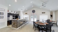 Located in Kingsport, Tennessee, this single-story space blends rustic charm with modern design throughout its thoughtfully arranged layout. The main living area immediately draws attention with its soaring vaulted ceilings, recessed lighting, and a dramatic floor-to-ceiling stacked stone fireplace with a wood mantel — a true centerpiece of the room. Comfortable seating surrounds a stylish coffee table, making this an inviting gathering space. Adjacent to the living area, an open-concept kitchen and bar area features dark cabinetry, a light countertop surface, and a reclaimed wood-paneled island with bar stool seating. French doors flood the space with natural light and provide direct access to the outdoors. A dedicated dining and activity area offers round table seating alongside decorative arrow wall accents and a workspace station tucked along the windowed wall. Throughout the space, neutral carpet flooring, white walls, and ceiling fans contribute to a clean, airy atmosphere. Glass-paned French doors connect distinct zones, maintaining an open feel while defining each area. The cohesive rustic-modern aesthetic is reinforced through natural textures, warm tones, and carefully chosen décor, creating a welcoming and functional environment well-suited for gathering and socializing.