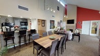 This single-floor community clubhouse is designed to foster comfort and social connection, featuring dramatic vaulted ceilings that lend a grand, open feel to the entire space. The main gathering area flows seamlessly between distinct zones, including a spacious dining area anchored by a large wood-tone table surrounded by upholstered chairs, and a relaxed lounge area furnished with a plush sofa, loveseat, and accent chairs arranged around a fireplace. Track lighting runs throughout the ceiling, providing warm, directional illumination that complements the neutral beige walls and carpeted floors. A bold red accent wall adds a striking architectural focal point, while framed artwork is thoughtfully displayed throughout, enhancing the welcoming ambiance. A kitchenette with a range, counter seating, and bar stools offers functional support for gatherings and events. Multiple seating vignettes create intimate conversation areas, including a cozy nook with rich brown accent chairs and a wall-mounted flat-screen TV. Large windows fitted with blinds line the perimeter, allowing generous natural light to fill the space while maintaining privacy. Lush indoor plants are positioned throughout, adding a refreshing, organic element. The layout is open and navigable, with clearly defined areas suited for dining, lounging, and socializing.
