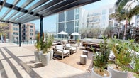 This expansive outdoor amenity space is designed for resort-style living within an urban environment. The centerpiece is a large, sparkling swimming pool flanked by a separate spa, both surrounded by smooth light-colored tile decking. A row of teak-framed chaise lounges with white cushions lines the poolside, offering ample space for sunbathing. Glass panel railings provide unobstructed sightlines, while the surrounding multi-story residential building creates a sheltered, courtyard-like atmosphere.

Adjacent to the pool area, a generously sized courtyard deck combines warm hardwood planking with stone-paved walkways. This zone is thoughtfully arranged with multiple seating vignettes, featuring modern outdoor sofas and armchairs with white cushions, concrete side tables, and dining tables with bench seating. White market umbrellas provide shade throughout. Tall, mature olive trees and tropical palms in large white planters are positioned throughout, adding natural texture and greenery to the contemporary aesthetic.

A partially covered pergola structure anchors one end of the courtyard, extending the usable space in varying weather. The overall design balances clean, modern architecture with organic landscaping elements, creating a welcoming and functional outdoor retreat with open sky views and an airy, California-inspired ambiance.