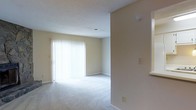 This two-floor residence offers a straightforward and functional layout designed to maximize comfort and usability. The living area, visible in the image, is finished with plush wall-to-wall carpeting in a neutral tone, contributing to a warm and quiet atmosphere. Large sliding glass doors fitted with vertical blinds serve as a prominent feature of the room, allowing an abundance of natural light to fill the space while also providing direct access to an outdoor area. The walls are painted in a soft, neutral beige, complementing the light carpeting and creating a cohesive, understated aesthetic throughout. A textured ceiling adds subtle architectural character to the room. The open sightlines between areas suggest a practical flow between living spaces. A smoke detector is visible, and standard electrical outlets are positioned conveniently along the walls. The two-floor configuration of the residence provides a natural separation between living and private spaces, a layout well-suited for both individuals and households seeking defined zones within the home. Overall, the space presents a clean, move-in-ready environment with an emphasis on natural light and neutral design.