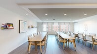 Located at 1112 First St in Alexandria, Virginia, this thoughtfully designed activity and meeting room offers a welcoming, functional environment suited for group gatherings, programming, and collaborative activities. The space is contained on a single floor and provides a comfortable yet spacious feel throughout.

The room features a clean, modern aesthetic with crisp white walls, a subtly tray-detailed ceiling with recessed lighting, and warm wood-tone flooring that runs the length of the space. Multiple long rectangular tables with mobile, upholstered chairs are arranged to accommodate group activities, and the layout can be easily reconfigured to suit various needs.

Large windows along one wall admit generous natural light and frame views of the surrounding Alexandria cityscape, lending an open, airy quality to the interior. Woven roller shades provide light control while maintaining the room's bright character.

Along one end of the room, a built-in kitchenette with gray cabinetry, a sink, countertop workspace, and a full-size refrigerator provides practical utility for events and programming. Organized shelving displays supplies and materials, and a doorway connects to an adjacent corridor, enhancing accessibility and flow.

Decorative touches—including framed artwork, a wall-mounted display shelf with colorful items, and hanging aprons—add warmth and personality, making this a functional yet inviting space for community engagement.