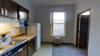 This single-floor space offers a straightforward and functional layout designed for practical everyday living. The kitchen area is equipped with essential appliances, including a full-size refrigerator and a dishwasher, complemented by wooden cabinetry and a light-toned countertop that provides ample workspace. An over-the-range microwave adds convenience to the cooking area. Adjacent to the kitchen, the space opens into a dining or living area distinguished by a large, double-hung window that draws in natural light and offers views of the surrounding urban streetscape. Beneath the window sits a classic cast-iron radiator, a hallmark of traditional building construction that provides reliable, efficient heating. The flooring transitions from resilient vinyl in the kitchen zone to carpet in the adjoining area, delineating the functional spaces. The walls are finished in a neutral gray tone, lending a clean and understated ambiance throughout. The overall layout is compact yet well-organized, making efficient use of the available space while maintaining a comfortable and livable atmosphere suited to urban residential living.
