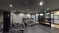 This fitness center is a thoughtfully designed, single-floor facility offering a comprehensive range of workout equipment to support both cardio and strength training. The space is finished with durable dark rubber flooring throughout, providing a safe and functional surface for exercise. Recessed lighting set within a drop tile ceiling illuminates the room evenly, creating a focused and energizing atmosphere.

The cardio zone features multiple True-brand treadmills and an elliptical trainer, each equipped with individual digital display screens. The strength training area includes a multi-function cable machine, a dedicated free weight rack stocked with a range of dumbbells, and adjustable weight benches for versatile workouts. Large wall mirrors along one side of the room visually expand the space and allow users to monitor their form during exercise.

A standout design feature is a large-scale, sepia-toned mural depicting a bustling street scene—likely inspired by a historic city district—spanning an entire wall and adding a distinctive artistic character to the space. Entry to the fitness center is through sleek black-framed glass doors, with a vending machine conveniently located nearby. The adjacent area, visible through the glass doors, suggests access to additional amenities. A posted fitness center guidelines notice and clearly marked exit signage complete the professional, well-organized environment.