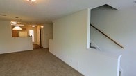 This two-floor residence offers a straightforward and functional layout designed for comfortable everyday living. The main level showcases a spacious living area finished with wall-to-wall carpeting in a warm neutral tone, providing a soft and inviting foundation throughout the space. The walls are painted in a crisp, light off-white, contributing to a bright and airy atmosphere.

A partially open staircase with a wooden handrail is nestled into one corner of the main level, providing access to the upper floor while maintaining a sense of openness in the living area. A low half-wall partition subtly defines the transition between the living space and the staircase area, adding a modest architectural detail without closing off the layout.

The hallway visible toward the rear of the main level leads toward the entry of the home, where a window allows natural light to filter in. The corridor connects the various functional areas of the main floor, offering a clear and accessible flow throughout the residence. The textured ceiling is consistent throughout, and the overall interior presents a clean, move-in ready condition with a simple, neutral palette.