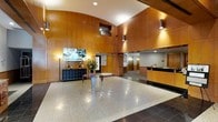 Located at 101 Summer Street in Stamford, Connecticut, this professional commercial lobby makes a strong first impression with its sophisticated and welcoming design. The space is defined by soaring ceilings that lend an expansive, airy quality, complemented by sweeping curved wood-paneled walls that add warmth and architectural distinction. Polished terrazzo flooring reflects the carefully considered lighting scheme, which combines recessed ceiling fixtures with sleek wall-mounted sconces to create a bright yet refined ambiance. A dedicated reception desk, finished in matching warm wood tones, is prominently positioned to greet and assist visitors upon entry. A central display table serves as a focal point in the open floor area, while decorative elements including floral arrangements and curated accent pieces contribute to a professional yet inviting atmosphere. A large flat-screen display mounted on the paneled wall provides an additional communication touchpoint. Elevator access is visible toward the rear of the lobby, ensuring smooth circulation throughout the building. The overall design balances corporate functionality with an elevated aesthetic, making this lobby well-suited as the front-facing hub of a professional office environment in downtown Stamford.