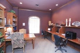 This single-floor home office space exudes a sophisticated, warm ambiance defined by its deep plum and rich burgundy wall tones, which are elegantly offset by natural wood trim, crown molding, and warm carpeted flooring throughout. The room is anchored by a substantial central wood table surrounded by upholstered floral-patterned chairs, offering a comfortable setting for meetings or collaborative work. A set of classic French doors with glass pane inserts connects the office to an adjacent bright living area, allowing natural light to filter in and creating a seamless sense of flow between spaces. A prominent arched window along one wall draws in additional daylight and frames a view of surrounding greenery. Built-in wood shelving and cabinetry line multiple walls, providing generous storage and display space for books, decorative items, and collectibles. A dedicated workstation area features a built-in countertop desk with ample surface area, accommodating office equipment including a multifunction printer. Recessed ceiling lighting complements the natural light to maintain a well-lit, productive atmosphere. The cohesive use of warm wood tones throughout the cabinetry, trim, and furniture ties the room together, lending the space a refined yet approachable character well-suited for both professional work and personal use.