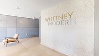 The lobby of Whitney Modern, located at 703 Whitney Avenue in New Haven, CT, serves as the welcoming gateway to this contemporary residential building. The space is defined by a refined modern aesthetic that blends architectural boldness with carefully curated design details. A dramatic feature wall adorned with a geometric gold-and-white faceted pattern prominently displays the "Whitney Modern" signage in mixed gold and metallic lettering, immediately establishing the building's identity and design sensibility upon entry. Large-format light stone tile flooring runs throughout the space, lending a clean, seamless foundation that enhances the sense of openness. Floor-to-ceiling glass walls and dark-framed sliding glass doors flood the lobby with abundant natural light while offering clear views of the surrounding streetscape, creating a strong visual connection between the interior and exterior. A textured concrete panel wall adds an industrial yet sophisticated contrast, providing architectural depth and visual interest. The seating area is appointed with mid-century modern leather arm chairs in warm caramel tones, paired with a glass-top coffee table and accented by fresh floral arrangements, contributing a welcoming and stylish atmosphere. Overhead, a sculptural brass sputnik chandelier adds a refined focal point. A resident mailbox alcove is discreetly integrated near the entryway, ensuring functional convenience within the elegant setting.