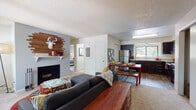 This single-floor residence offers a well-organized layout that balances comfort and functionality. The open-concept living area serves as the heart of the home, anchored by a fireplace with a rustic wood-panel accent wall and a white mantle, adding a distinctive focal point to the space. Large windows along the front wall invite natural light and provide views of the surrounding greenery. The living area transitions seamlessly into the dining space, which features hardwood-style flooring and direct access to the kitchen. The kitchen is equipped with dark espresso cabinetry, stainless steel appliances including a full-size refrigerator, a double sink positioned beneath a window, and ample counter space. The unit includes two carpeted bedrooms, each with sliding closet doors and windows that bring in natural light. One bedroom offers a more serene, neutral palette, while the second provides a relaxed, casual feel. The bathroom features a combined tub and shower with sliding glass doors, a dark-cabinet vanity with a white countertop, and tile flooring. Throughout the residence, textured ceilings and gray-toned walls create a cohesive and modern aesthetic.