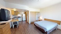 This single-floor studio unit offers an open-concept layout that efficiently combines the living, sleeping, and kitchen areas into one cohesive space. The unit features warm wood-tone laminate flooring throughout, lending a clean and consistent look to the interior.

The kitchen is positioned along one wall and is equipped with light wood cabinetry, a full-size refrigerator, an electric range, and a sink with ample counter space. A small dining table with seating for two is conveniently situated just adjacent to the kitchen, providing a dedicated area for meals.

The sleeping area occupies the opposite end of the unit and includes a full-size bed with a nightstand and lamp, creating a comfortable resting space. A sliding door closet nearby offers practical storage solutions for clothing and personal items.

The unit's open design maximizes the sense of space, with neutral white walls and recessed ceiling elements contributing to a bright and airy atmosphere. The straightforward, functional layout makes this studio a practical option for individuals seeking an all-in-one living space with essential amenities readily at hand.