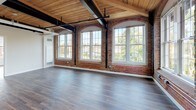Located at 83 Mechanic St in Attleboro, Massachusetts, this thoughtfully renovated loft-style unit showcases a striking blend of historic industrial character and contemporary design. The open living area is defined by exposed red brick walls, soaring wood-plank ceilings with bold black steel beams, and rich hardwood flooring that runs throughout the space. Oversized arched and grid-pane windows flood the interior with abundant natural light while framing views of the surrounding greenery.

The kitchen is well-appointed with crisp white shaker cabinetry, stone-look countertops, and a full suite of stainless steel appliances, including a side-by-side refrigerator with ice and water dispenser, a range, an over-the-range microwave, and a dishwasher. The clean, modern finishes of the kitchen contrast beautifully with the raw, historic textures found throughout the rest of the unit.

A dedicated bedroom area benefits from its own set of large windows with tree-lined views, continuing the warm wood ceiling and hardwood floor aesthetic. The bathroom is finished in a clean, neutral palette with large-format tile flooring, a vanity with ample storage, and a bright, airy feel. Together, these spaces create a cohesive unit that honors its mill-building heritage while delivering a polished, move-in-ready living experience.