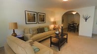 This single-floor residence features a comfortable living room designed with a warm, neutral color palette throughout. The space is anchored by plush, tan upholstered seating, including a sofa and matching armchair, both accented with teal and patterned throw pillows that add subtle pops of color. A dark wood coffee table with a lower shelf serves as a functional centerpiece, styled with decorative candle holders and floral accents.

The room is finished with wall-to-wall carpeting in a light beige tone, contributing to the overall warmth of the space. A textured ceiling is fitted with a flush-mount ceiling light, providing even overhead illumination. Two framed art pieces adorn one wall, adding a decorative touch to the otherwise clean, simple surfaces.

Natural light enters through large windows dressed with vertical blinds and golden-yellow drape panels, creating a bright and airy atmosphere. A window-mounted air conditioning unit ensures year-round comfort. The overall layout is practical and inviting, making efficient use of the available space while maintaining a tidy, livable ambiance.