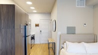 This thoughtfully designed single-floor studio presents a clean, modern aesthetic with a highly functional open-concept layout. Light hardwood flooring runs continuously throughout, lending warmth and visual cohesion to the entire space. Recessed ceiling lighting provides even, bright illumination across the unit.

The kitchen is positioned along one wall and is fully equipped with stainless steel appliances, including a refrigerator, range, and over-the-range microwave. Warm wood-toned cabinetry provides ample storage, complemented by a sleek countertop workspace. The kitchen flows seamlessly into the main living and sleeping area, which is framed by neutral, light-toned walls that enhance the sense of openness.

The sleeping area accommodates a full-sized bed with an upholstered headboard and is anchored by a wall-mounted flat-screen television. A wire-frame shelving unit serves as a flexible open wardrobe and storage solution. Near the entry, a keypad-secured front door is flanked by a wall-mounted mirror and a slim entryway bench, creating a practical and welcoming arrival experience.

A standout feature is the glass door leading to a private balcony, accompanied by a sidelight window that draws in natural light and provides outdoor views. The unit also includes a wall-mounted thermostat and electrical panel, ensuring all essential systems are accessible within the space.