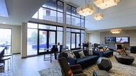 This contemporary single-floor lounge space makes an immediate impression with its dramatic double-height ceilings and expansive floor-to-ceiling window walls that flood the interior with natural light. The towering glazed facade frames views of the surrounding exterior, including a stone-clad building façade and an outdoor patio or pool area, creating a seamless connection between indoors and out. Large glass doors provide direct access to the outdoor space, enhancing the open, airy feel of the interior. The lounge is thoughtfully furnished with a mix of navy blue sofas and armchairs accented with geometric throw pillows in warm orange and neutral tones, arranged atop a patterned area rug to create an inviting conversational seating area. High-top bistro tables with sleek black chairs are positioned near the windows, offering a casual spot for socializing or working. Overhead, a series of large, square pendant light fixtures with warm-toned shades provide elegant ambient lighting that complements the natural light. The flooring transitions between light hardwood and tile, adding visual texture throughout. The overall ambiance is modern, sophisticated, and welcoming, making it an ideal gathering space for socializing or relaxation.