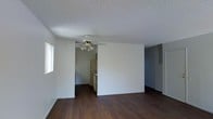 This single-story space offers a straightforward and functional layout designed for comfortable everyday living. The main living area is finished with rich, dark hardwood-style flooring that runs throughout, providing a clean and cohesive look underfoot. A ceiling fan with a built-in light fixture is centrally mounted, ensuring both air circulation and ambient lighting across the open space. The walls are painted in a crisp, neutral white, and the textured popcorn ceiling adds a classic touch overhead. Natural light filters in through a window along one wall, brightening the interior without overwhelming it. An open passage connects the living area directly to the kitchen, which is outfitted with white cabinetry and white appliances, including a refrigerator and range, offering a tidy and efficient cooking space. To the right of the main living area, a built-in shelving or closet unit provides dedicated storage, adding practical utility to the layout. The overall design is clean, minimal, and move-in ready, making it a functional space well-suited for a variety of living arrangements.