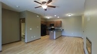 This single-floor space is designed with an open-concept layout that seamlessly connects the living area and kitchen, creating a bright and functional environment. The kitchen is a standout feature, equipped with dark black appliances including a refrigerator, stove, and built-in microwave. Rich, warm-toned wood cabinetry lines the upper and lower walls, offering ample storage, while a peninsula with a dark countertop provides additional workspace and casual seating potential. Light hardwood flooring extends throughout the main living area, adding warmth and a sense of continuity to the space. Recessed lighting in the kitchen is complemented by flush-mount ceiling fixtures, and a ceiling fan with an integrated light serves the living area, ensuring comfortable airflow and illumination. The neutral, taupe-toned walls provide a versatile backdrop suitable for a variety of décor styles. A glimpse into an adjacent bathroom is visible, hinting at additional functional spaces within the unit. Overall, the layout is practical and inviting, well-suited for everyday living.