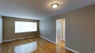 This single-floor space presents a straightforward and practical layout designed for comfortable living. The main room is finished with warm-toned hardwood floors that reflect natural light, contributing to a bright and inviting atmosphere. The walls are painted in a neutral gray-taupe tone, providing a versatile backdrop suitable for a variety of interior styles. A large window along one wall allows ample natural light to fill the space, enhancing its open feel. A flush-mount ceiling light fixture provides additional illumination. White baseboards and door trim offer clean contrast against the neutral walls, adding a crisp, finished look throughout. An open doorway connects this main room to an adjacent space, visible in the background, which shares the same hardwood flooring and light-toned walls, suggesting a consistent design language across the unit. The overall ambiance is calm and understated, making it a functional and move-in ready space with a neutral palette that accommodates personalization. The absence of clutter and the clean finishes give the space a well-maintained and orderly character.