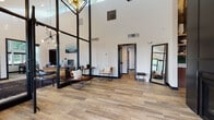 This single-floor commercial space blends contemporary design with warm, inviting details to create a highly functional and visually striking environment. The main reception and common area immediately impresses with dramatic soaring ceilings and bold floor-to-ceiling black-framed glass partitions that define individual work zones while maintaining an open, airy feel. A statement Sputnik chandelier anchors the space overhead, complementing the crisp white walls and rich wood-look tile flooring that runs throughout. The entrance foyer features a sleek black fireplace set against a floor-to-ceiling surround, adding a refined focal point near the front glass doors. Private offices and meeting rooms branch off the central corridor, each accessible through black-trimmed doorways that maintain the cohesive design aesthetic. The kitchen and lounge area is a standout, featuring a central island with a herringbone-tiled base, white quartz countertops, and a deep farmhouse sink. Open wood shelving lines a dramatic dark accent wall adorned with large-scale botanical artwork, while exposed ductwork adds an industrial touch. A communal dining table with black chairs provides a collaborative gathering space. Abundant natural light filters in through multiple windows, enhancing the modern, professional ambiance throughout.