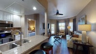 This single-floor residence offers a thoughtfully designed layout that balances comfort and functionality. The open-concept living area serves as the heart of the home, featuring rich dark hardwood flooring, a ceiling fan, and large windows dressed with elegant floor-length draping curtains that invite abundant natural light. The living room is anchored by a plush neutral sofa, a dark wood coffee table, and a wall-mounted flat-screen television, creating a warm and inviting atmosphere.

The kitchen flows seamlessly from the living space, showcasing granite countertops, crisp white cabinetry with modern hardware, and stainless steel appliances including a full-size refrigerator. A breakfast bar with seating provides a casual dining option.

The bedroom is a serene retreat, appointed with soft carpeting, warm neutral tones, and vaulted ceilings that enhance the sense of space. Large framed artwork adorns the wall above the bed, and the room leads directly into a spacious walk-in closet. The bathroom is finished with granite countertops, white cabinetry, a large vanity mirror, and decorative wall accents, offering both practicality and style.

Throughout the residence, a cohesive warm color palette of taupe, cream, and gold tones creates an inviting and refined ambiance.