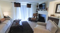 This single-floor space offers a warm and inviting atmosphere centered around a standout living area. The room is anchored by a classic white fireplace with dark tile surround, which serves as a natural focal point and adds a touch of elegance to the space. Above the fireplace, built-in shelving provides both functional storage and display space for decorative accents. A wall-mounted flat-screen television is seamlessly integrated into the entertainment setup, making the area ideal for relaxation and hosting. Large windows dressed with layered curtains — featuring rich brown and white tones — allow abundant natural light to pour in, creating a bright and airy feel throughout. The living area is thoughtfully furnished with a neutral sofa, a tufted accent chair, and a dark coffee table resting atop a plush area rug, all contributing to a cohesive and comfortable aesthetic. The neutral carpeted flooring ties the room together, complementing the warm brown tones found throughout the décor. Overall, the space strikes a balance between comfort and style, making it a welcoming environment suited for everyday living.