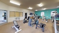 This fitness center in Bowling Green, Kentucky offers a well-equipped, single-floor workout facility designed to support a full range of exercise routines. The open layout is finished with neutral carpeted flooring and bright white walls accented by a bold teal feature wall, creating an energizing and welcoming atmosphere. Flush-mounted fluorescent lighting ensures the space is consistently and evenly lit throughout.

The cardio section is well stocked, featuring multiple treadmills, an elliptical trainer, a stationary bike, and a rowing machine, providing ample options for cardiovascular workouts. A dedicated dumbbell rack with a full range of free weights and accompanying flat benches supports strength and resistance training. Several cable and weight machines line the perimeter, further expanding the variety of strength training options available.

A full-wall mirror along one side of the room enhances the sense of space and allows members to monitor their form during workouts. Multiple wall-mounted televisions provide entertainment throughout the session. Arched windows along the exterior walls allow natural light to filter into the space, contributing to an open and airy feel.

A small utility area near the rear of the facility includes a sink and cabinetry, offering practical convenience for members. A water fountain is also accessible within the space. The overall design is functional and straightforward, prioritizing ease of use and a comfortable workout environment.