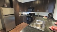 This single-floor residence offers a thoughtfully designed living space with a strong emphasis on modern aesthetics and practical functionality. The kitchen stands out as a central highlight, showcasing rich dark espresso cabinetry that extends from floor to ceiling, providing generous storage throughout. Granite-style countertops in deep, earthy tones complement the cabinetry and add a sense of refined character to the space. The kitchen is equipped with a full suite of black appliances, including a gas range, over-the-range microwave, and a stainless steel refrigerator, creating a cohesive and contemporary look. A double stainless steel sink with a modern faucet anchors the countertop workspace, making it ideal for meal preparation. The warm hardwood-style flooring running through the kitchen adds a natural contrast to the darker upper elements, enhancing the overall warmth of the interior. The layout is designed to maximize efficiency, with ample counter space and a well-organized work triangle between the sink, stove, and refrigerator. The overall ambiance is sleek and inviting, blending dark tones with warm wood finishes to create a kitchen environment that is both stylish and highly functional.