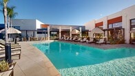Located at 41461 Buecking Drive in Temecula, California, this generously sized outdoor amenity space offers a resort-inspired pool experience spanning a substantial footprint. The centerpiece is a large, sparkling swimming pool with crystal-clear blue water, framed by a wide, open-air deck finished in neutral-toned pavers that reflect the abundant Southern California sunshine. Rows of sling-style chaise lounges and striped cabana umbrellas are thoughtfully arranged along the poolside, providing ample space for relaxation and leisure. Accent water features add a dynamic visual element to the pool's edge. The surrounding landscape incorporates drought-tolerant plantings and tall palm trees that reinforce the warm, California atmosphere. A modern clubhouse structure with floor-to-ceiling glass panels and clean geometric lines anchors one end of the deck, offering an elegant architectural backdrop. The space is enclosed by a contemporary multi-story residential building featuring a bold palette of white, teal, and red exterior panels. Mountain views are visible on the horizon, enhancing the open, airy quality of the environment. The overall design balances modern aesthetics with functional outdoor living, creating a vibrant communal gathering space well-suited to the sunny Temecula climate.