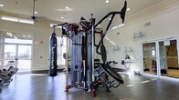 This single-floor fitness center is thoughtfully designed to provide a comprehensive workout environment for users of all fitness levels. The space is anchored by a large, professional-grade multi-functional cable and strength training machine, offering a wide variety of resistance exercises from a single station. A heavy-duty punching bag is suspended nearby, catering to those interested in boxing and martial arts conditioning. A well-stocked dumbbell rack lines one wall, providing a full range of free weights for strength training. The flooring consists of dark hardwood, lending a clean and polished aesthetic to the space. Natural light filters in through large windows, keeping the atmosphere bright and energizing, while recessed ceiling lighting ensures consistent illumination throughout. The walls are adorned with decorative bicycle artwork, adding a sporty and modern visual character to the room. High windows along the upper perimeter further enhance the sense of openness. The overall ambiance is both functional and inviting, striking a balance between a professional gym setting and a welcoming community fitness space.