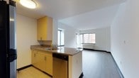 This single-floor residence offers a thoughtfully designed open-plan layout that seamlessly connects the kitchen and living areas. The kitchen is appointed with warm yellow cabinetry, granite-style countertops, a stainless steel sink, and a built-in dishwasher. A peninsula island extends the counter space and serves as a natural divider between the kitchen and the living area, making it ideal for both meal preparation and casual dining. The flooring transitions from tile in the kitchen to wood-style flooring in the living space, adding a sense of definition while maintaining an open and airy feel. Large windows in the living area allow generous amounts of natural light to flood the interior, providing views of the surrounding urban streetscape, including glimpses of neighboring brick buildings. The clean, white walls and bright ceilings contribute to the overall sense of openness throughout the space. A baseboard heating unit is visible beneath the windows, indicating a well-considered approach to climate comfort. The overall ambiance is clean, functional, and modern, making it a versatile space well-suited for everyday living.