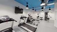 This fitness center offers a thoughtfully designed workout space that accommodates a variety of exercise needs. The room's most distinctive architectural feature is its vibrant blue ceiling, which contrasts sharply with the clean white walls and light gray flooring, creating an energetic yet focused atmosphere. Exposed ductwork and industrial-style black pendant lights hang from the ceiling, providing ample illumination throughout the space.

The cardio section is well-stocked with Spirit-brand treadmills and an elliptical trainer, arranged in an organized row along one side of the room. A wall-mounted flat-screen television is positioned for easy viewing during cardio workouts. On the opposite end of the space, a multi-station cable machine offers a comprehensive range of strength training options, complemented by a dedicated free weights area featuring a double-tiered dumbbell rack stocked with varying weights and accompanying flat benches. A recumbent bike provides an additional low-impact cardio option. The space also includes a water cooler and waste receptacle for convenience. Multiple access points via white-paneled doors connect to adjacent areas, and the open floor plan ensures comfortable movement between equipment stations.