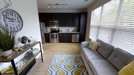 This single-floor space showcases an open-concept design that seamlessly integrates the kitchen and living area into one cohesive, functional environment. The kitchen is outfitted with full-height dark espresso cabinetry that extends to the ceiling, providing generous storage while creating a bold visual contrast against the light-toned countertops and pale hardwood-style flooring that runs throughout the space. Stainless steel appliances, including a refrigerator, electric range, microwave, and dishwasher, lend a polished, modern aesthetic to the kitchen. A single-basin sink is positioned along the cabinetry wall, keeping the layout efficient and streamlined. The living area flows naturally from the kitchen, offering comfortable space for seating and relaxation. A gray sofa anchors the room, complemented by a patterned area rug in blue and gold tones that adds warmth and visual interest underfoot. A slim console table provides additional surface space along one wall. A large window dressed with horizontal blinds allows natural light to filter into the space, brightening the interior and offering a glimpse of greenery outside. Track lighting overhead ensures even, adjustable illumination throughout.