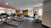 Located at 1520 University Blvd NE in Albuquerque, New Mexico, this fitness center is a well-equipped, single-floor facility designed to support a comprehensive workout experience. The space is divided into distinct zones, each tailored to a specific type of exercise.

The cardio area is prominently outfitted with a row of True-brand treadmills and elliptical trainers, arranged to maximize space efficiency. A large wall mirror expands the visual depth of this zone, while recessed lighting illuminates the room evenly. A ceiling fan adds comfort during high-intensity workouts.

Adjacent to the cardio area, a dedicated strength training zone features a multi-station cable machine and a functional trainer, ideal for resistance and weight-based exercises. An anatomical reference chart is posted on the wall, providing guidance for users. A bold orange accent wall, decorated with a heartbeat graphic, energizes this section of the facility and adds a motivational visual element.

A stationary bike provides an additional low-impact cardio option. The rubber flooring throughout the space offers durability and safety underfoot. Natural light enters through a window near the entry door, which leads to an outdoor area. Fitness area rules are posted prominently, ensuring proper use of the facility. Overall, the space delivers a functional and well-organized environment suited for a full range of fitness activities.