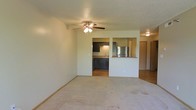 This single-floor residence offers a practical and comfortable layout designed for everyday living. The living area is spacious and carpeted, providing a warm and inviting atmosphere. A ceiling fan with an integrated light fixture keeps the space well-ventilated and well-lit. The textured ceiling adds a subtle dimensional quality to the room.

The kitchen is accessible directly from the living area and features dark cabinetry that contrasts nicely with the light-toned countertops. A stainless steel refrigerator anchors the kitchen, complemented by a sink positioned along the cabinetry wall. A pass-through counter opening connects the kitchen to the living space, creating an open, communicative flow between the two areas — ideal for entertaining or casual dining.

The flooring transitions from carpet in the living area to hard flooring in the kitchen, offering both comfort and practicality where needed. Neutral wall tones throughout contribute to a clean, understated aesthetic that allows for versatile interior styling. A hallway visible from the living area suggests additional rooms beyond, completing this well-organized single-floor layout.