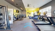 Located at 450 Allure Cir in Whiteland, Indiana, this fitness center is a well-appointed, single-floor exercise facility designed to support a comprehensive workout experience. The space is defined by a bold, energizing aesthetic — bold yellow accent walls contrast sharply with dark gray tones, while motivational signage such as "DON'T QUIT!" reinforces an atmosphere of determination and focus. Ceiling fans and linear LED lighting run throughout, keeping the environment comfortable and well-illuminated. Large floor-to-ceiling windows and a garage-style roll-up door flood the space with natural light and offer open views of the surrounding community, creating an airy, connected feel. Full-length mirrors along one wall visually expand the room while serving a functional purpose for form-checking during workouts. The exercise floor is finished with durable rubber flooring accented by teal directional chevron graphics. Equipment is thoughtfully arranged throughout the space and includes a robust free weight section with a full dumbbell rack, a weight bench, and a cable machine. Cardio options are equally impressive, with multiple treadmills, elliptical trainers, and spin/cycling bikes — many equipped with individual screens. Mounted flat-screen televisions add to the entertainment options available during workouts. The overall design balances high energy and functionality, making it a motivating environment for users of all fitness levels.