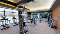 This fitness center is designed to provide a comprehensive workout experience within a single, open-plan floor. The space features a thoughtfully arranged selection of equipment, including treadmills, ellipticals, cable machines, and a multi-station strength trainer from Hoist, catering to a full range of fitness needs. Large floor-to-ceiling windows line one side of the room, flooding the interior with natural light and offering views of the surrounding outdoor landscape. The ceiling showcases an industrial-style design with exposed ventilation ductwork, complemented by recessed lighting and a vaulted section that adds verticality to the space. A deep teal accent wall provides a bold, energizing contrast to the neutral tones of the patterned carpet flooring. Wall-mounted mirrors on one side effectively expand the sense of space and allow users to monitor their form during workouts. Mounted televisions are positioned throughout for added entertainment. The overall ambiance is modern, clean, and well-maintained, making it a functional and inviting environment for fitness enthusiasts of all levels.