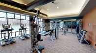This fitness center is designed to provide a comprehensive workout experience within a single, open-plan floor. The space features a thoughtfully arranged selection of equipment, including treadmills, ellipticals, cable machines, and a multi-station strength trainer from Hoist, catering to a full range of fitness needs. Large floor-to-ceiling windows line one side of the room, flooding the interior with natural light and offering views of the surrounding outdoor landscape. The ceiling showcases an industrial-style design with exposed ventilation ductwork, complemented by recessed lighting and a vaulted section that adds verticality to the space. A deep teal accent wall provides a bold, energizing contrast to the neutral tones of the patterned carpet flooring. Wall-mounted mirrors on one side effectively expand the sense of space and allow users to monitor their form during workouts. Mounted televisions are positioned throughout for added entertainment. The overall ambiance is modern, clean, and well-maintained, making it a functional and inviting environment for fitness enthusiasts of all levels.