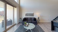 This contemporary two-story loft-style residence is defined by its dramatic double-height ceilings on the main floor, which create an expansive, airy atmosphere throughout the living area. Large clerestory windows flood the space with natural light, while a full-width sliding glass door opens onto a private balcony offering sweeping city skyline views — a standout feature of the living room. The flooring on the main level is durable wood-look planking that runs throughout, complementing the modern aesthetic. The kitchen is outfitted with stainless steel appliances, a mosaic tile backsplash, white upper cabinetry, and a dark island with bar seating and pendant lighting overhead. A dedicated home office on the main level features carpeted flooring and a ceiling fan, providing a quiet workspace. An open staircase with a sleek railing leads to the upper level, where the bedroom suite offers plush carpeting, recessed lighting, and direct access to a well-appointed en suite bathroom featuring dark granite countertops, an undermount sink, and wood-look tile flooring. A second full bathroom mirrors the same refined finishes, complete with a shower/tub combination. The overall design balances industrial-loft character with modern residential comfort.
