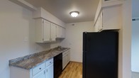The kitchen in this single-floor space is thoughtfully designed to maximize functionality within a compact footprint. White cabinetry lines the upper and lower walls, providing ample storage while contributing to a clean, bright aesthetic. The countertops feature a granite-style surface with subtle earth-tone patterning, offering a durable and visually appealing workspace. Black appliances, including a full-size refrigerator and dishwasher, create a striking contrast against the lighter cabinetry and walls. A stainless steel sink is positioned along the counter, complemented by under-cabinet storage and a functional layout that allows for efficient movement throughout the space. Warm hardwood-style flooring runs throughout, adding a touch of warmth to the overall design. Overhead, a flush-mount ceiling light provides consistent illumination, while HVAC vents integrated into the ceiling ensure year-round climate comfort. The neutral wall tones and understated design elements give the kitchen a versatile, move-in-ready appeal suitable for a variety of personal styles.