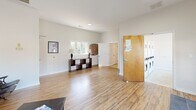 Located at 648 Holly Ave in Winston-Salem, NC, this single-floor space offers a well-organized layout comprising two living areas, a dedicated laundry room, and a full bathroom. The interior is finished with light wood-look flooring throughout the main areas and neutral white walls, creating a bright and welcoming atmosphere.

The primary living area features soaring vaulted ceilings that lend an open, airy feel, complemented by large multi-pane windows that flood the room with natural light and provide views of the surrounding greenery. A secondary living area connects seamlessly, also benefiting from generous window placement and high ceilings.

The laundry room is equipped with multiple side-by-side washer and dryer units, accessible via a solid wood door, and includes a folding counter workspace beside a large picture window overlooking the parking area — a practical and functional setup. The bathroom is designed with accessibility in mind, featuring stainless steel grab bars positioned around the toilet and a window for natural ventilation. The space is finished with tile flooring consistent with its functional purpose.

Overall, the space presents a clean, functional, and community-oriented environment with thoughtful accessibility features, ample natural light, and a cohesive neutral design palette throughout.