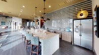 This contemporary single-floor community space is thoughtfully designed to blend comfort and sophistication. The open-concept layout flows seamlessly from the fully appointed kitchen and bar area into generous lounge and seating zones, creating an inviting environment for social gatherings and relaxation.

The kitchen area is a standout feature, showcasing a striking textured tile accent wall with horizontal striped detailing, complemented by sleek wood cabinetry, granite and stone countertops, and stainless steel appliances. A large central island with terrazzo-style countertops and stylish leopard-print upholstered bar seating serves as a natural gathering point, illuminated by a series of bold black pendant lights.

The main lounge area is anchored by curved contemporary sofas in warm neutrals and accented with vibrant orange and terracotta throw pillows atop a bold chevron rug. Stone-clad columns add an architectural grounding element, while an artistic chandelier adds a dramatic focal point overhead.

Floor-to-ceiling windows along the main living area flood the space with natural light and offer unobstructed views of the outdoor pool and surrounding landscaping, visually connecting the interior to the resort-style amenity area. Dark hardwood-style flooring runs throughout, tying the various zones together with a cohesive, upscale aesthetic.