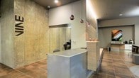 This single-floor space presents a refined industrial aesthetic, anchored by a sleek reception area that serves as the focal point upon entry. The design balances raw and polished elements seamlessly — expansive exposed concrete walls provide an industrial backdrop, while a clean, modern reception desk finished in cool grey offers a striking contrast. A wood-paneled accent wall adds warmth and texture to the space, complementing the wood-look tile flooring that runs throughout.

Overhead, a trio of copper-toned pendant lights hang at varying heights above the reception desk, casting a warm ambient glow that softens the industrial character of the space. Recessed ceiling lighting provides additional illumination, ensuring the area remains bright and welcoming. A glass partition behind the reception desk offers a subtle visual connection to an adjacent workspace or office area, maintaining an open feel while delineating functional zones. Small decorative accents on the reception counter add a personalized touch. The overall ambiance is professional yet inviting, making it well-suited for a business-facing environment such as a salon, clinic, or boutique office.