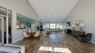 This expansive single-floor gathering space is defined by its dramatic vaulted ceilings that rise steeply overhead, creating an airy, open atmosphere throughout. Recessed lighting and an elegant chandelier provide warm illumination, complementing the rich hardwood-style flooring that spans the entire space.

The main lounge area is generously proportioned, featuring multiple seating arrangements including plush sofas with accent pillows, a velvet accent chair, and a decorative area rug anchoring the central sitting zone. A tile-surround fireplace serves as a focal point on one wall, with a wall-mounted flat-screen television mounted above. A carved sideboard and curated artwork add refined detail to the space.

Large windows along the rear wall frame picturesque views of an outdoor pool, gazebo, and landscaped grounds, flooding the interior with natural light. French doors provide direct access to the outdoor area. A mirrored wall and bistro-style seating nook offer additional gathering options near the entrance.

A compact kitchenette, outfitted with gray shaker cabinetry, white countertops, a stainless steel refrigerator, and a sink, supports the space's function as a social or event venue. The neutral gray color palette and cohesive furnishings throughout contribute to a polished, welcoming ambiance.