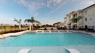 This single-story community clubhouse showcases a thoughtfully designed coastal aesthetic throughout its welcoming interior and outdoor amenities. The grand lobby greets visitors with soaring ceilings, exposed decorative beams, and an abundance of natural light pouring in through oversized windows. Herringbone-patterned hardwood flooring extends across the open main area, adding warmth and visual texture to the space. A dramatic circular chandelier serves as a centerpiece above a round accent table, anchoring the room with refined elegance. Teal leather wingback chairs and coastal-inspired artwork, including abstract blue canvases and decorative wall medallions, reinforce the property's nautical color palette. A sliding barn door leads to an adjoining lounge equipped with a fireplace and flat-screen television, offering a comfortable retreat. Rich mahogany double-entry doors open to the exterior on multiple sides, seamlessly connecting the indoors to the outdoors. Outside, a generously sized resort-style swimming pool takes center stage, surrounded by a clean white deck, palm trees, and wrought iron fencing. Views of the surrounding residential community and open sky complete the outdoor experience, creating a bright and inviting atmosphere year-round.