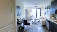 This well-appointed single-floor residence offers a thoughtfully designed open-concept layout that seamlessly blends living and dining areas into one cohesive and functional space. The interior is anchored by rich hardwood-style flooring that runs throughout, providing a warm and contemporary foundation for the décor.

The living area is generously sized, featuring a plush neutral sofa, a sleek mirrored coffee table, and a patterned area rug that adds texture and visual interest. A ceiling fan with a modern profile provides both comfort and style overhead. Large windows and a full glass sliding door flood the space with natural light, creating a bright and airy ambiance while offering views of the surrounding outdoors.

Adjacent to the living area, an open shelving unit serves as a subtle room divider, leading into a dining nook furnished with a glass-top table and geometric-style chairs. A stylish bar cart near the entryway adds a sophisticated, functional touch, making it ideal for entertaining.

The entryway is highlighted by a distinctive oversized decorative medallion that sets a personalized and design-forward tone from the moment you enter. Overall, the space presents a modern, curated aesthetic with attention to detail throughout, balancing comfort and elegance in a compact yet efficient single-level floor plan.