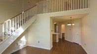 This two-story home offers a thoughtfully designed interior that balances openness with functional living spaces. The main level immediately impresses with rich hardwood flooring that extends throughout, warm neutral wall tones, and crisp white trim and baseboards that lend a clean, classic aesthetic. A striking open staircase with white balusters and a natural wood handrail rises to the upper level, where an open railing balcony overlook adds vertical dimension and architectural interest to the space below. The soaring double-height ceiling above the main living area creates a generous sense of volume and airiness. Toward the back of the main level, a peninsula-style kitchen counter with warm wood cabinetry provides a natural transition into the kitchen and dining areas. An elegant chandelier with multiple light fixtures anchors the dining space, adding a refined touch to the overall ambiance. The kitchen connects seamlessly to adjacent rooms, supporting an easy flow for everyday living and entertaining. The upper level, visible from below through the open balcony railing, provides additional private living quarters. Throughout the home, the combination of hardwood floors, neutral finishes, and carefully chosen lighting fixtures creates a warm, welcoming atmosphere that is both timeless and move-in ready.