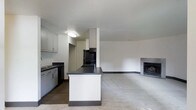 This single-floor space showcases an open-concept layout that seamlessly connects the kitchen and living areas. The kitchen is well-appointed with gray cabinetry, dark countertops, and black appliances, including a range and dishwasher. A central island with a dark countertop and white paneling provides additional workspace and serves as a natural divider between the kitchen and the adjoining living area. Flush-mount ceiling lighting illuminates the kitchen, while a dedicated alcove houses a tall upper cabinet above the range. The living area extends generously from the kitchen, featuring neutral carpet flooring and a tiled fireplace along the far wall, adding a focal point to the space. The open layout creates a sense of spaciousness, with clean white walls and a neutral color palette throughout contributing to a bright, minimalist aesthetic. A hallway visible from the kitchen suggests additional rooms beyond the main living area. Overall, the space balances functional design with understated modern style.