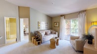 This single-floor residence is thoughtfully designed with a warm, neutral color palette that creates a calm and inviting atmosphere throughout. The living room serves as the heart of the space, featuring plush carpeting, soft sage-green and beige walls, and abundant natural light streaming in through a large sliding glass door that opens onto a private balcony. The balcony offers lush greenery views, including what appears to be tropical foliage, enhancing the serene ambiance. A decorative grid mirror anchors one wall of the living room, complementing the understated artwork and layered décor accents, including woven side tables and sculptural decorative pieces that add texture and character. The living area transitions smoothly into an adjacent room — visible through an open doorway — which appears to serve as a dining or study space, with its own natural light source and clean design. The overall layout is open and airy, with a cohesive design language that emphasizes comfort and simplicity. Light-toned furnishings, soft drapery, and carefully chosen accessories contribute to a refined yet approachable aesthetic, making this a well-appointed and functional living environment.