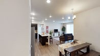 This single-floor residence is thoughtfully designed with an open-concept layout that seamlessly connects the kitchen, dining, and living areas. The kitchen is a standout feature, showcasing floor-to-ceiling dark espresso cabinetry, crisp white countertops, and a full suite of stainless steel appliances including a side-by-side refrigerator with water dispenser, electric range, and built-in microwave. A central island with bar seating adds both prep space and casual dining functionality. Light wood-look flooring runs throughout the main living areas, complementing the neutral white walls and recessed lighting overhead. The open dining area is illuminated by an elegant chandelier, while the living room offers a comfortable, welcoming atmosphere with ample natural light from nearby windows. The primary bedroom is generously sized with plush carpeting, a dedicated walk-in closet, and a private en suite bathroom featuring dark vanity cabinetry and a sliding glass shower enclosure. A secondary bedroom with a window overlooking the exterior provides a bright, cozy retreat. The second full bathroom includes a tub-shower combination. Throughout the residence, the design balances modern finishes with a warm, livable aesthetic, creating a cohesive and functional single-level home.