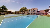 Located at Shady Oaks Manor in Fort Worth, Texas, this outdoor pickleball facility offers a well-appointed recreational space designed for active community living. The complex features multiple side-by-side pickleball courts, each finished in a striking two-tone blue and green sport surface with crisp white boundary lines and regulation-height nets. The courts are fully enclosed by sturdy black chain-link fencing, ensuring safe and defined play areas throughout. A mature shade tree anchors the space, providing a natural focal point and relief from the Texas sun. The surrounding landscape includes maintained green lawns, ornamental shrubs, and flowering crape myrtles that add vibrant color to the setting. A low decorative retaining wall lines one edge of the complex, adding structure to the grounds. The facility is set against a backdrop of two-story residential buildings with neutral siding and pitched roofs, creating a cohesive community atmosphere. Wide open skies and abundant natural light make this an inviting venue for both casual play and more competitive matches. Walkway access along the perimeter ensures easy entry and exit. The overall setting strikes a balance between functional athletic infrastructure and a pleasant, park-like environment suited for year-round outdoor activity in the Fort Worth community.